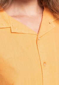 Camicia a maniche corte con colletto di un vivace arancione, realizzata in un tessuto strutturato con due dettagli di bottone, caratterizzata da un collo aperto e una finitura pulita.