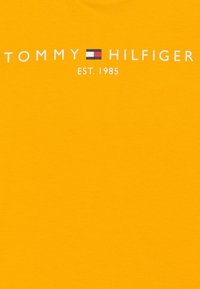 Żółta bawełniana koszulka z logo „TOMMY HILFIGER” z czerwonym, białym i niebieskim akcentem w formie bloku oraz napisem „EST. 1985” poniżej.
