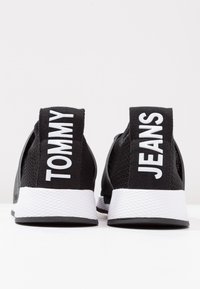 Czarne buty sportowe z siateczkową cholewką, białymi podeszwami i wyraźnym brandingiem na zakładkach piętowych, na których widnieje napis "TOMMY" i "JEANS". Teksturowane wykończenie.