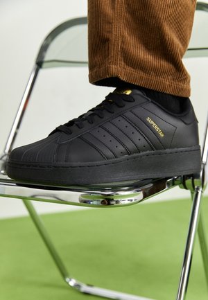 Zwarte Adidas Superstar sneaker met gouden merklogo, gedragen met bruine corduroy broek, rustend op een chromen inklapbare stoel op een groene vloer.