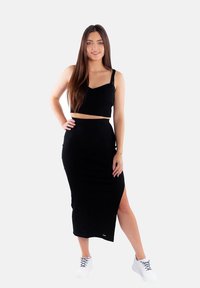 Czarny prążkowany crop top na cienkich ramiączkach w zestawieniu z dopasowaną czarną spódnicą midi z bocznym rozcięciem, noszony z białymi trampkami.