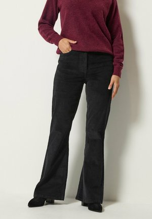 Personne portant un pantalon large en velours côtelé noir, des chaussures noires et un pull bordeaux à manches longues, debout avec une main dans la poche.