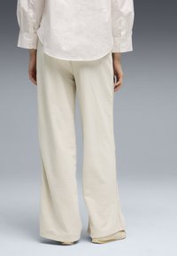 Personne portant un pantalon ample couleur crème, une chemise blanche à manches longues et des chaussures beiges, debout devant un fond dégradé gris.