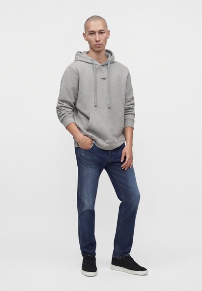 Jeune homme debout, portant un sweat à capuche gris avec poche avant, un jean bleu et des baskets noires avec semelles blanches, sur un fond blanc uni.
