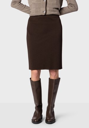 Femme portant une jupe marron foncé jusqu'aux genoux, des bottes marron foncé montant jusqu'aux genoux et une veste boutonnée à motifs, les mains sur les hanches.