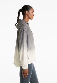 Giubbotto leggero con cappuccio in gradazione dal grigio chiaro al bianco. Presenta polsini elastici e tasche laterali, progettato per l'abbigliamento attivo.