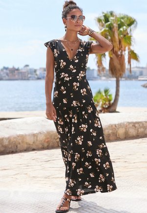 Frau trägt ein schwarzes Maxikleid mit Blumenmuster und Sonnenbrille, geht auf einem steinernen Weg am Meer entlang, umgeben von Palmen bei sonnigem Wetter.
