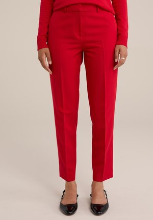 Vrouw die op maat gemaakte rode broek en zwarte slingback hakken van lakleer draagt, staand tegen een effen lichte achtergrond.