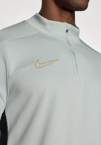 Ανοιχτό μπλε φούτερ της Nike με γιακά και φερμουάρ μέχρι το στήθος, με χρυσό κεντημένο λογότυπο. Ομαλή υφή και εφαρμοστό σχέδιο με μαύρες πλευρικές λεπτομέρειες.