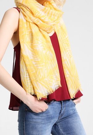 Femme portant un foulard jaune à motifs sur un haut rouge sans manches et un jean bleu, les mains reposant dans les poches avant.