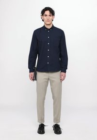 Navy button-up shirt with long sleeves, two chest pockets, paired with light beige tapered trousers and black lace-up shoes. Minimalist design.