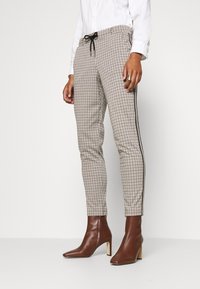 Pantalon beige à carreaux avec taille élastique, rayures latérales et coupe ajustée, associé à des bottines marron et une chemise blanche.