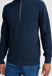 Tamno plavi pulover s teksturiranim pletenim dizajnom, polu-zip prednjim dijelom i rebrastim rukavima i rubom, u kombinaciji s plavim trapericama.
