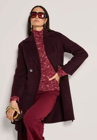 Vrouw in een oversized donker bordeauxrode jas, bordeaux gebreide trui, bijpassende broek, grote rode zonnebril, met een bruine clutch met gouden handvat.