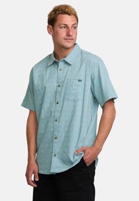 Camisa de manga corta de botones en azul claro con un estampado de diamantes, bolsillo frontal y botones marrones. Fabricada con un tejido ligero y transpirable.