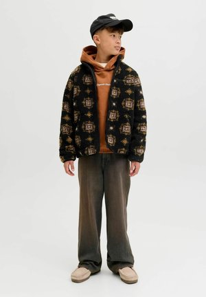 Zwarte fleecejack met een patronen en bruine accenten, gedragen over een oranje hoodie. Losse grijze broek en beige schoenen maken de outfit compleet.