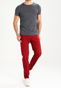 Hombre vestido con una camiseta ajustada de color gris oscuro, pantalones rojos brillantes y zapatillas blancas caminando sobre un fondo blanco.