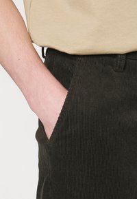 Pantalon en velours côtelé vert foncé, avec une poche latérale et une texture côtelée visible. La boucle de ceinture est également présente. Chemise beige visible en haut.