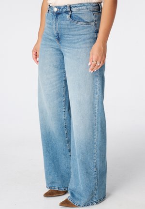 Persoon die lichtblauwe wijde jeans en bruine puntige schoenen draagt, met ontspannen handen langs het lichaam tegen een witte achtergrond.