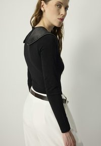 Mujer vestida con una blusa negra de manga larga y gran cuello, y pantalones blancos de tiro alto con un cinturón negro, mirando por encima del hombro.
