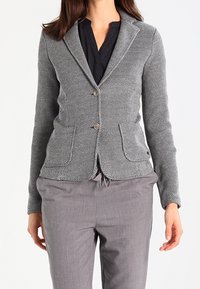 Blazer grigio con texture a spina di pesce, a singolo petto, due bottoni e due tasche frontali, abbinato a pantaloni grigi.