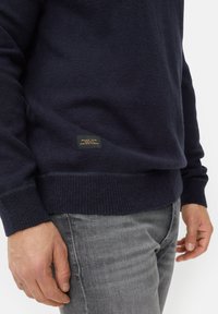 Navy crewneck sweater made of soft knit fabric; features ribbed cuffs and hem, with a small label tag near the bottom showcasing branding.