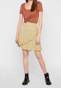 Hnědý top s krátkým rukávem a uzlíčkem doplněný žlutou kvetinovou mini sukní s volánkovým lemem, podtržený černými kotníkovými botami.