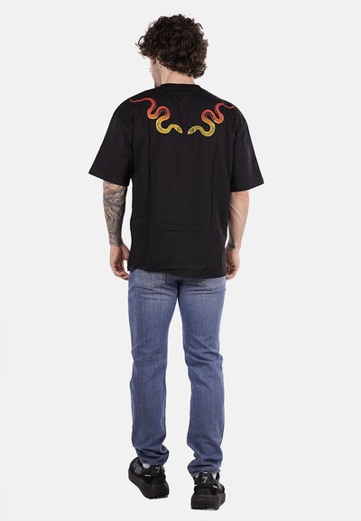 T-shirt nera con una vestibilità ampia, caratterizzata da ricami di serpenti rossi e gialli sulla schiena. Abbinata a jeans blu e sneakers nere.