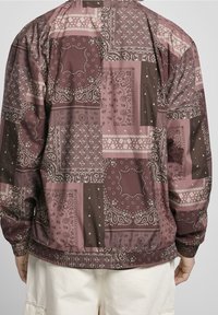 Bakifrån av en person som bär en maroon- och mauve-färgad jacka med patchworkbandana-mönster och ljusbeige byxor.