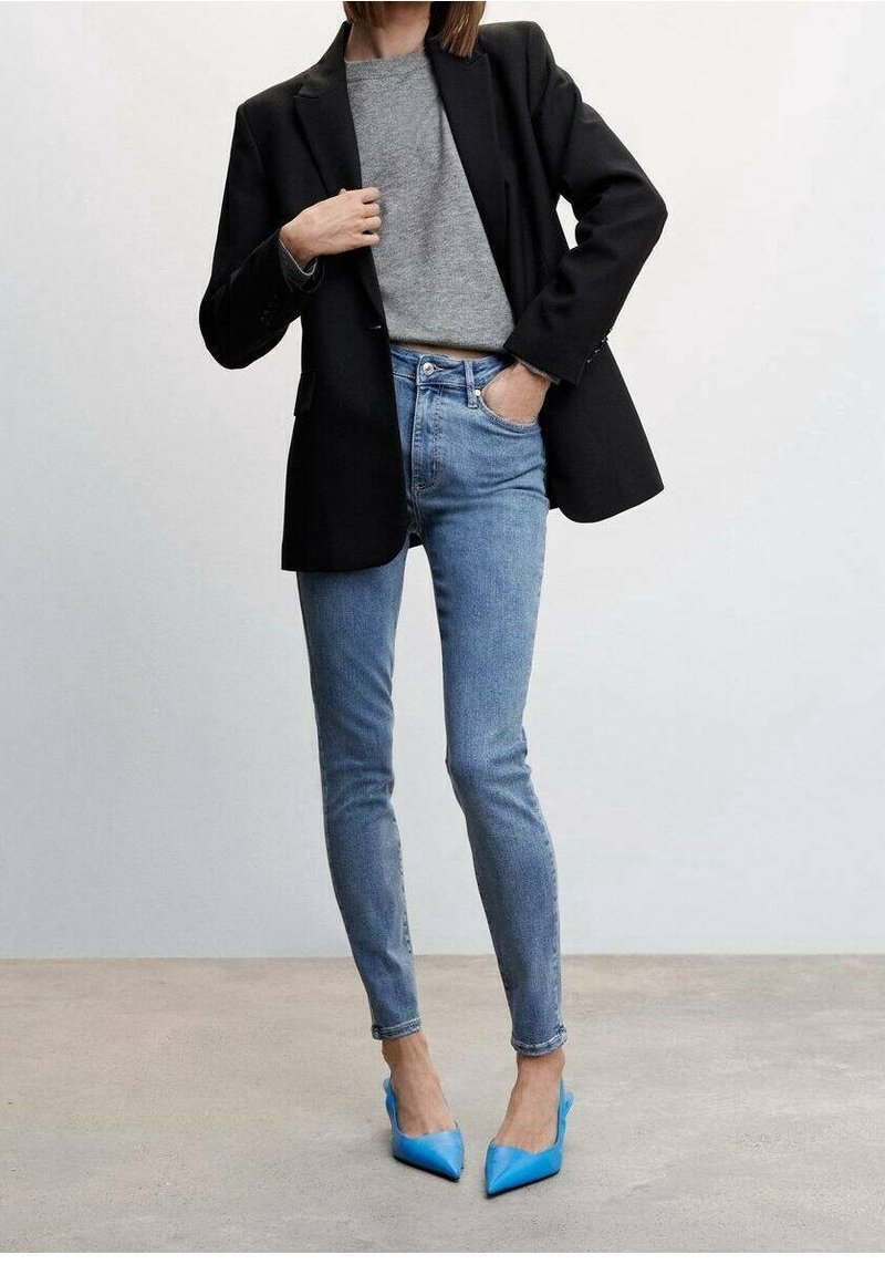 Femme portant un blazer noir, un pull gris, un jean skinny bleu et des escarpins pointus bleu vif, debout sur un sol en béton.