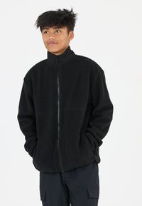 Veste en polaire noire avec un col haut et une fermeture éclair intégrale, dotée de deux poches avant et de poignets côtelés. Texture douce et coupe oversize.