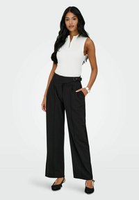 Bílý žebrovaný top bez rukávů se zipem vpředu, kombinovaný s černými širokými kalhotami s vysokým pasem, které mají boční pásek a kapsy.