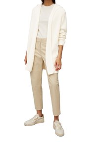 Žena oblečená v bílém pleteném cardiganě bez zapínání, off-white tričku, béžových kalhotách po kotníky a krémových teniskách stojící na bílém pozadí.