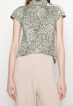 Femme portant un chemisier à manches courtes et col montant imprimé léopard et un pantalon en maille côtelée beige clair, debout, les bras détendus le long du corps.