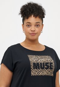 Sort T-shirt med korte ærmer, der har et grafik med tan zebra-striber og teksten "MUSE" i fede, blokbogstaver. Blødt bomuldsstof.