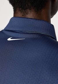 Granatowa koszulka polo o teksturowym wzorze, z białym logo Nike na tylnym kołnierzu oraz standardowym złożonym kołnierzykiem.