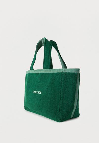 Πράσινη τσάντα ώμου από πετσέτα, με δύο λαβές, ανοιχτό πράσινο περίγραμμα και λευκή επιγραφή «Versace» στην μπροστινή πλευρά.