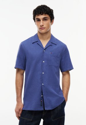 Jeune homme aux cheveux courts et bouclés portant une chemise bleue à manches courtes avec une poche poitrine et un jean foncé sur fond blanc.