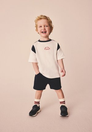 Junges Kind mit blondem Haar steht, lächelt, trägt ein weißes und schwarzes T-Shirt, schwarze Shorts, gestreifte Socken und schwarze Turnschuhe.