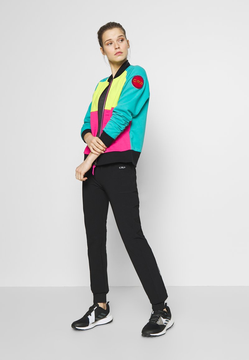 Farbige Jacke mit Reißverschluss in Türkis-, Zitronen- und Pinkpaneelen, schwarzem Besatz; kombiniert mit schwarzen Hosen und schwarzen Sportschuhen.