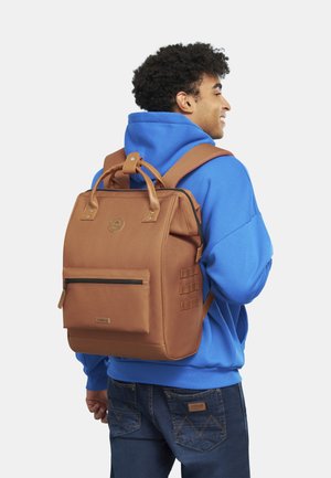 Sac à dos en toile marron avec des accents en cuir, poignées supérieures et bretelles réglables. Comprend une poche zippée à l'avant et des détails en sangle sur les côtés.