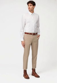 Camicia bianca con bottoni, pantaloni beige, cintura in pelle marrone e stivaletti marroni. Tessuto liscio, vestibilità sartoriale e design minimalista.