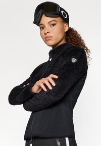 Must puhvaga must fleece-jakk, millel on tekstuuriga, koheline välimus ja siledad külgpaanid. Omab randmel embleemi ja kõrget kaelust.