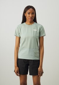 Tricou din bumbac de culoare verde deschis, cu mâneci scurte, având o buzunar frontal și o etichetă mică cu marca pe piept. Purtați-l cu pantaloni scurți negri.