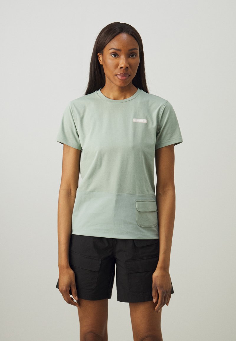 Tricou din bumbac de culoare verde deschis, cu mâneci scurte, având o buzunar frontal și o etichetă mică cu marca pe piept. Purtați-l cu pantaloni scurți negri.