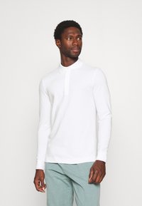 Långärmad vit polo skjorta med krage, med en subtil logotyp på bröstet, tillverkad av ett mjukt tyg, ihop med ljusblå byxor.