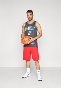 Szürke kosárlabda mez "DETROIT" felirattal és "2" számmal, piros nadrággal párosítva. Fehér tornacipő, fekete fejpánt, és egy kosárlabda a kezében.