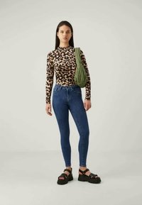 Dugi rukav, majica s leopard printom, plave uske traperice, zelena torba za rame i crne platforme. Glatka tekstura s uskim dizajnom.