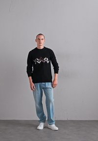 Man som står mot en grå vägg iförd en svart sweater med ett zigzagmönster, ljusblå jeans och vita sneakers.