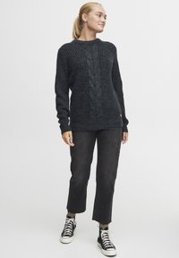 Donkergrijze gebreide trui met een kabelpatroon in het midden, ronde halslijn en geribbelde boorden, gecombineerd met zwarte cropped broek en sneakers.